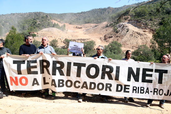 Membres de les plataformes Almatret Net i la Ribera Digna en l'acció per denunciar el projecte de l'abocador de residus de Riba-roja i al fons les màquines que treballen en els terrenys | ACN